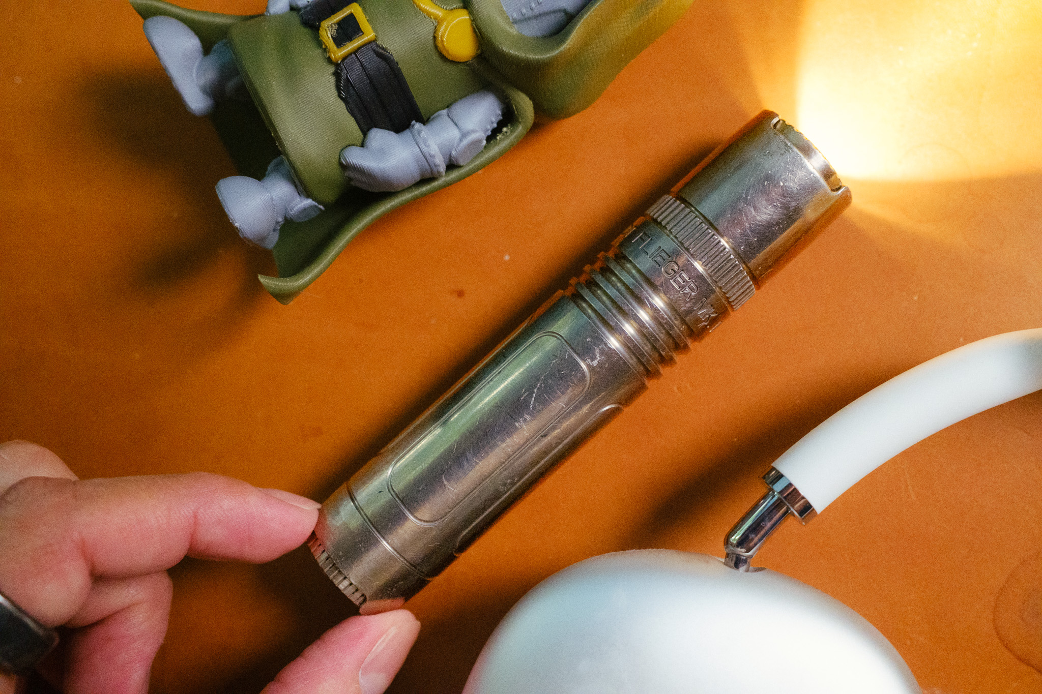 The flashlight, on, being held by the end with thumb and pointer finger, next to a dr. doom toy and AirPods Max for scale